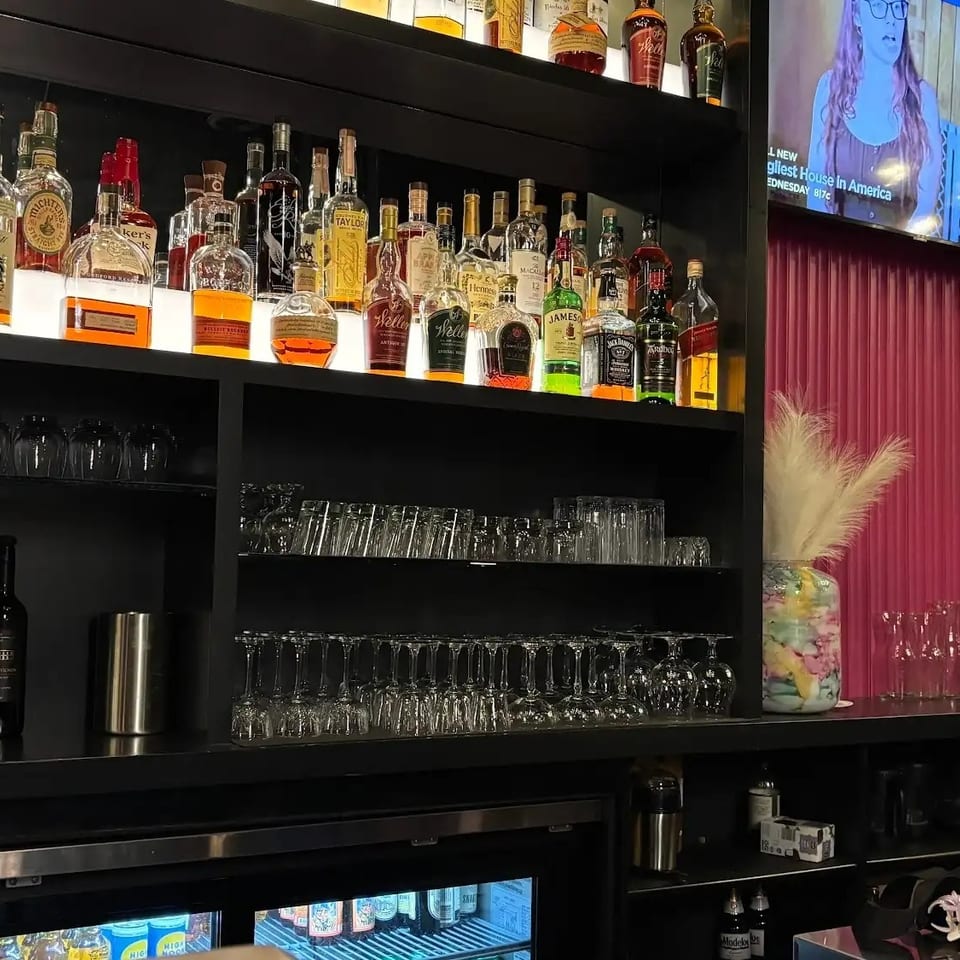Restaurant bar with illuminated shelves of liquor bottles, rows of glassware, a television, decorative flowers, and table lamp.